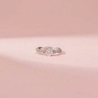 Front view of a 14KT white gold ring featuring a round-cut diamond in a criss-cross design, set against a pink background.
