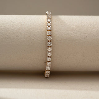Front view of a classic gold tennis bracelet featuring a continuous line of round brilliant gemstones in secure prong settings, displayed on a soft neutral cylindrical stand.