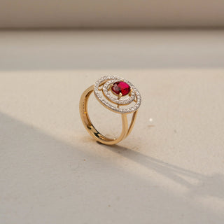 Angled view of a gemstone halo ring in 14 KT Yellow Gold, featuring an oval-cut red center stone encircled by concentric pavé-set diamond halos in an elevated open design, displayed against a beige backdrop.