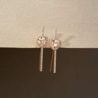 Side view of diamond drop earrings in 14 KT Rose Gold, featuring heart-inspired pavé diamond motifs with floating center diamonds and sleek linear diamond accents against a neutral backdrop.
