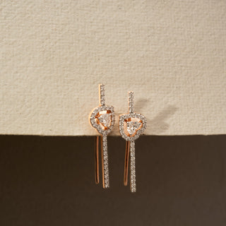Angled view of diamond drop earrings in 14 KT Rose Gold, featuring heart-inspired pavé diamond motifs with floating center diamonds and sleek linear diamond accents against a neutral backdrop.
