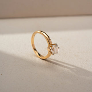 Angled view of a round solitaire engagement ring in 14 KT Yellow Gold, featuring a round lab-grown diamond in a classic six-prong setting on a polished gold band, displayed against a beige backdrop.