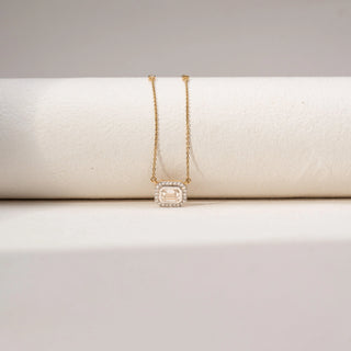 Front view of an emerald-cut lab-grown diamond pendant necklace in 18KT Yellow Gold, featuring a bezel-set emerald-cut centre stone with a sparkling diamond halo. Captured against a beige backdrop, the elegant design radiates brilliance.