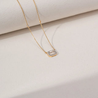 Angled view of an emerald-cut lab-grown diamond pendant necklace in 18KT Yellow Gold, featuring a bezel-set emerald-cut centre stone with a sparkling diamond halo. Captured against a beige backdrop, the elegant design radiates brilliance.