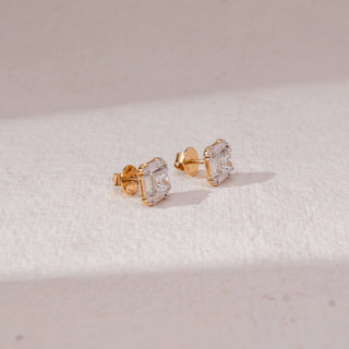 Angled view of square halo diamond stud earrings in yellow gold, featuring a centred princess-cut lab-grown diamond framed by baguette and round stones. Placed on a textured beige surface, the design casts a soft shadow.
