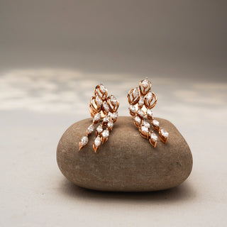 Front view of cascading diamond drop earrings in 14 KT Rose Gold, featuring marquise-cut diamonds in layered leaf-inspired motifs, displayed on a textured stone surface.
