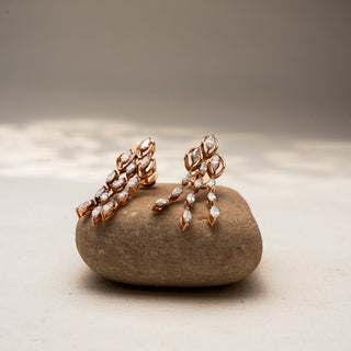 Angled view of cascading diamond drop earrings in 14 KT Rose Gold, featuring marquise-cut diamonds in layered leaf-inspired motifs, displayed on a textured stone surface.
