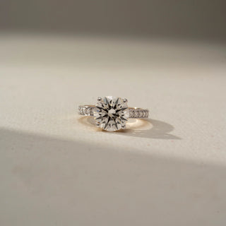 Front view of a solitaire diamond ring featuring a round brilliant-cut center stone with pavé diamond accents on the band, set in warm-toned gold and displayed in soft natural light.