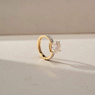 Angled view of a yellow gold solitaire ring featuring a large oval-cut diamond and a pavé-set band, displayed on a neutral surface with soft natural lighting.