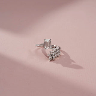 Top view of a white gold diamond ring featuring an emerald-cut center stone and a curved marquise diamond leaf motif with pavé accents, displayed on a soft pink background.