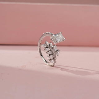 Side view of a white gold diamond ring featuring an emerald-cut center stone and a marquise diamond leaf cluster with pavé accents, displayed on a soft pink background.
