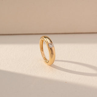 Side view of Flush Set Diamond Band Ring in 14KT Yellow Gold, featuring a single round lab grown diamond embedded in a smooth band. Set on a beige backdrop, the design highlights sleek minimal elegance.
