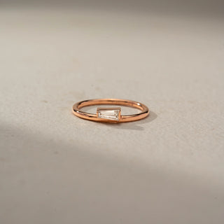 Front view of a baguette solitaire ring in 14 KT Rose Gold, featuring a horizontally set baguette-cut lab-grown diamond on a sleek minimalist band, displayed against a beige backdrop.