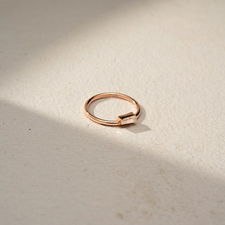 Side view of a baguette solitaire ring in 14 KT Rose Gold, featuring a horizontally set baguette-cut lab-grown diamond on a sleek minimalist band, displayed against a beige backdrop.