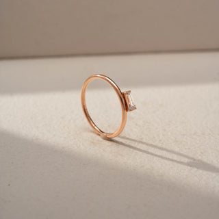 Angled view of a baguette solitaire ring in 14 KT Rose Gold, featuring a horizontally set baguette-cut lab-grown diamond on a sleek minimalist band, displayed against a beige backdrop.