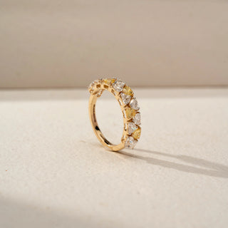 Angled view of a diamond eternity band in 14 KT Yellow Gold, featuring alternating heart-shaped yellow and white lab-grown diamonds in elevated shared-prong settings, displayed against a beige backdrop.