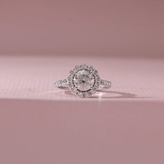 Front view of round diamond halo ring in white gold with a pavé band, set on a soft pink background, highlighting classic design and bright, multi-layered sparkle.
