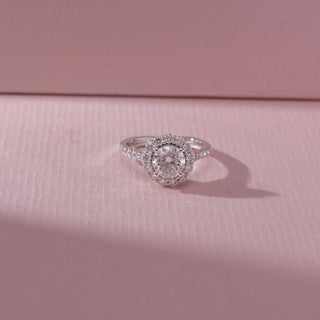 Top view of round diamond halo ring in white gold with a pavé band, set on a soft pink textured surface, highlighting classic symmetry and radiant, layered sparkle.