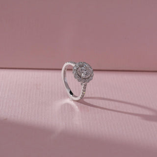 Angled view of round diamond halo ring in white gold with a pavé band, set on a soft pink textured background, highlighting delicate detailing and radiant sparkle.
