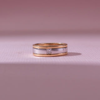 Front view of a two-tone gold band ring, featuring a centered princess-cut diamond set within a silver-toned stripe. Captured against a soft pink backdrop, the design reflects modern elegance and minimalism.
