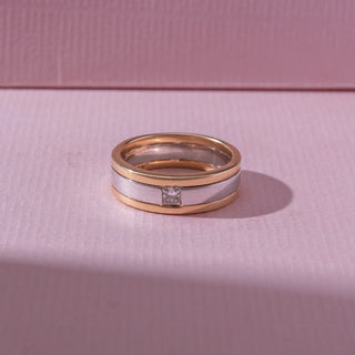 Top view of a two-tone gold band ring, featuring a centered princess-cut diamond set within a silver-toned stripe. Captured against a soft pink backdrop, the design reflects modern elegance and minimalism.
