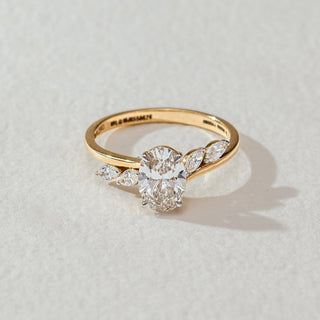 Top view of oval diamond ring in yellow gold, featuring a centred oval lab-grown diamond with marquise side stones on a curved band. Placed on a textured beige surface, the design casts a soft shadow.
