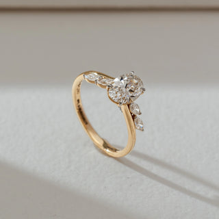 Side view of round diamond ring in yellow gold, featuring a centred round lab-grown diamond with marquise and pear side stones on a delicate band. Placed on a textured beige surface, the design casts a soft shadow.
