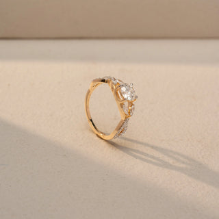 Angled view of a round diamond engagement ring in 14 KT Yellow Gold, featuring a round lab-grown diamond accented by marquise-shaped and pavé-set diamonds in a nature-inspired band, displayed against a beige backdrop.
