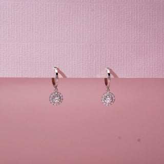Front view of drop diamond hoop earrings in white gold, featuring circular halo pendants with round lab-grown diamonds. Suspended against a soft pink backdrop, the design reflects delicate brilliance.
