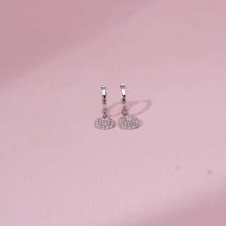 Front view of drop diamond hoop earrings in white gold, featuring circular halo pendants with round lab-grown diamonds. Placed on a soft pink surface, the design casts a gentle shadow.
