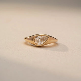 Front view of a pear-cut solitaire signet ring in 14 KT Yellow Gold, featuring a bezel-set pear-shaped lab-grown diamond in a sleek tapered band, displayed against a beige backdrop.