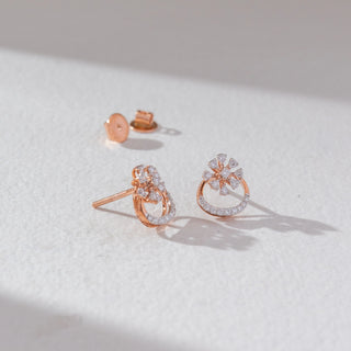 Angled view of floral diamond stud earrings in rose gold, featuring pear-shaped lab-grown diamonds in a blooming cluster above a curved diamond-studded base with visible screw backs. Set on a soft neutral surface.
