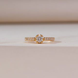 Front view of Modern Collet Pavé Diamond Ring in yellow gold, highlighting a round lab-grown diamond in a collet setting with pavé diamond accents along the band, displayed on a soft beige surface.
