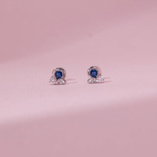 Front view of sapphire-style diamond stud earrings in white gold, featuring a central blue stone with round lab-grown diamonds below. Placed on a soft pink surface, the design reflects elegant contrast.
