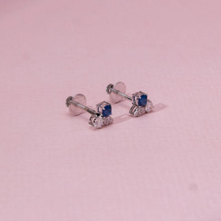 Angled view of sapphire-style diamond stud earrings in white gold, featuring a central blue stone with round lab-grown diamonds below. Placed on a soft pink surface, the design casts a soft shadow.
