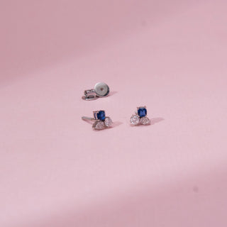 Top view of sapphire-style diamond stud earrings in white gold, featuring a central blue stone with round lab-grown diamonds below. Placed on a soft pink surface, the design casts a soft shadow.
