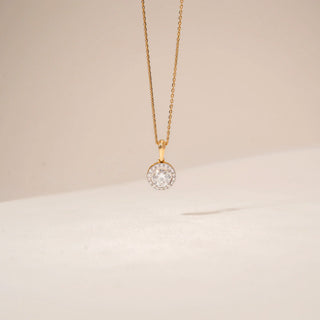 Front view of a halo diamond pendant in 14KT Yellow Gold, featuring a round centre diamond surrounded by smaller stones against a beige backdrop.