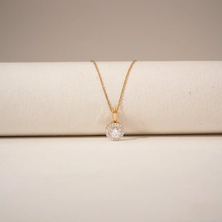 Close-up view of a halo diamond pendant in 14KT Yellow Gold, featuring a round centre diamond surrounded by smaller stones against a beige backdrop.