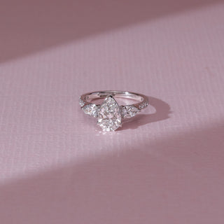 Top view of pear diamond ring in white gold, featuring a centred pear-shaped lab-grown diamond with matching side stones on a pavé band. Placed on a soft pink surface, the design casts a soft shadow.
