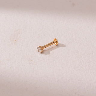 Angled view of a yellow gold nose pin, featuring a solitaire round diamond in a minimal prong setting. Captured against a soft beige backdrop, the design reflects delicate sparkle and understated elegance.
