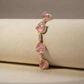 Close Up view of a pink gemstone bracelet in 14 KT Yellow Gold, featuring pear-shaped pink cabochon stones linked by diamond-set connectors and accented with pavé diamond swirls, displayed against a soft beige backdrop.
