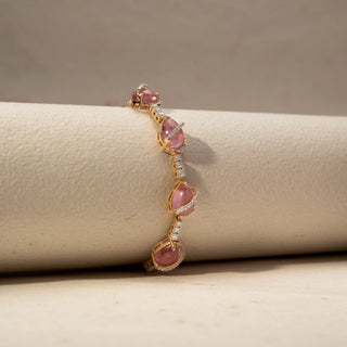 Side  view of a pink gemstone bracelet in 14 KT Yellow Gold, featuring pear-shaped pink cabochon stones linked by diamond-set connectors and accented with pavé diamond swirls, displayed against a soft beige backdrop.

