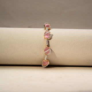 Front view of a pink gemstone bracelet in 14 KT Yellow Gold, featuring pear-shaped pink cabochon stones linked by diamond-set connectors and accented with pavé diamond swirls, displayed against a soft beige backdrop.
