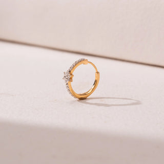 Side view of a gold nose ring, featuring pavé-set diamonds with a floral diamond accent at the center. Captured against a soft beige backdrop, the design reflects delicate sparkle and elegance.
