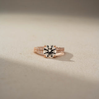 Front view of a round solitaire engagement ring in 14 KT Rose Gold, featuring a round lab-grown diamond in a four-prong setting with a triple-row pavé diamond band, displayed against a beige backdrop.