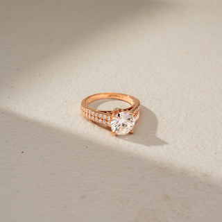 Top view of a round solitaire engagement ring in 14 KT Rose Gold, featuring a round lab-grown diamond in a four-prong setting with a triple-row pavé diamond band, displayed against a beige backdrop.