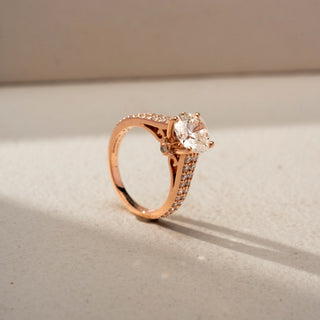 Angled view of a round solitaire engagement ring in 14 KT Rose Gold, featuring a round lab-grown diamond in a four-prong setting with a triple-row pavé diamond band, displayed against a beige backdrop.