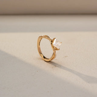 Angled view of a princess-cut solitaire engagement ring in 14 KT Yellow Gold, featuring a princess-cut lab-grown diamond set on a twisted pavé diamond band, displayed against a beige backdrop.