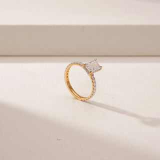 Angled view of a radiant  diamond ring in 14KT Yellow Gold, featuring a pavé-set band and four-prong centre stone against a beige backdrop.