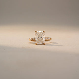 Front view of a radiant-cut diamond engagement ring in 14 KT Yellow Gold, featuring a radiant-cut lab-grown diamond with pavé-set diamonds along the band, displayed against a beige backdrop.